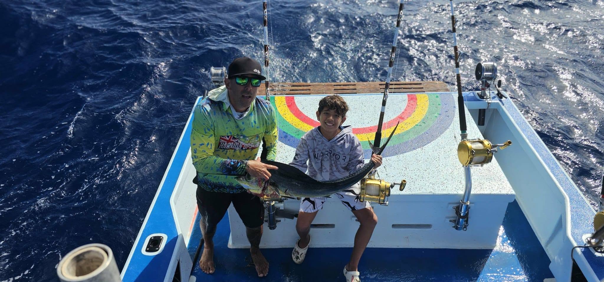 An adult and a child holding a mahi aboard Rainbow Runner at sea